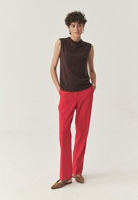 Rjava brezrokavnik, rdeče krojene hlače in rjave pointed flats. Model stoji z rokami v žepih, nevtralna ozadje.