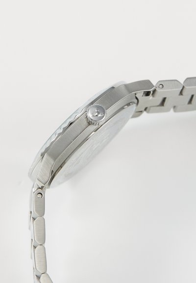 Relógio de aço inoxidável prateado com uma coroa texturizada, caixa octogonal e pulseira de elos com acabamentos polidos e escovados.