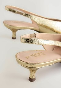 Los tacones slingback dorados presentan un acabado metálico texturizado, un tacón stiletto bajo y una plantilla rosa suave con un logotipo en relieve.