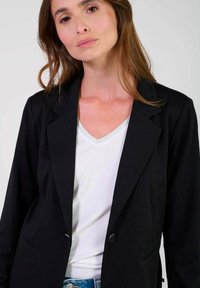 Svart kavaj med en strukturerad design, markerade slag, en knappstängning och sidofickor, bärs över en vit V-ringad topp.