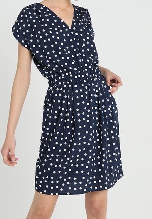 Femme portant une robe bleu marine longueur genou à pois blancs, manches courtes, col en V et taille élastique, debout devant un fond uni.