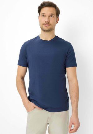 Man met een lichte huid en kort bruin haar, gekleed in een marineblauw T-shirt en beige broek, staand met één hand in de zak, kijkend naar boven.