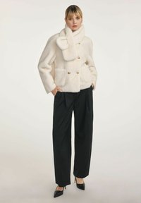 Kobieta w kremowym, puszystym płaszczu z dopasowanym szalikiem, w czarnych szerokich spodniach i czarnych szpilkach, stojąca z rękami w kieszeniach.