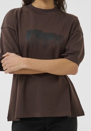 Osoba w luźnym, ciemnobrązowym T-shircie z czarnym, abstrakcyjnym wzorem przypominającym farbę na klatce piersiowej, z założonymi na sobie rękami na wysokości talii.