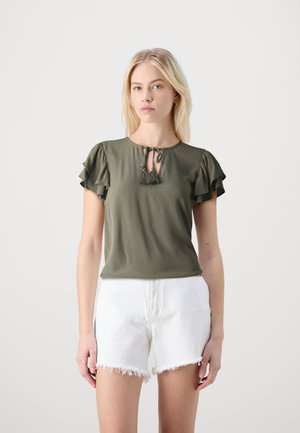 Olivengrønn topp med fløyelskort armer, nøkkelhullsutringning med knyting, kombinert med hvite shorts med frynsete hem. Glatt tekstur.