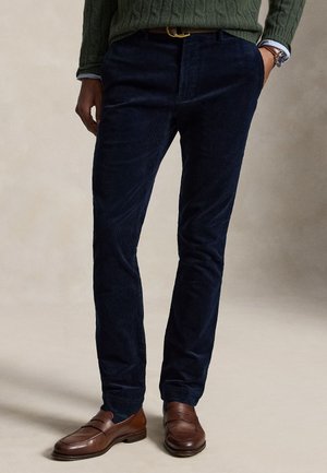 Homme portant un pantalon en velours côtelé bleu foncé, des mocassins en cuir marron, un pull vert et une montre, debout avec une main dans la poche.