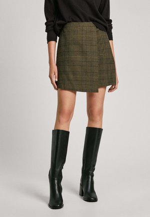 Falda de cuadros verde y amarilla con cintura alta y un diseño corto y acampanado, combinada con botas de cuero negras hasta la rodilla.