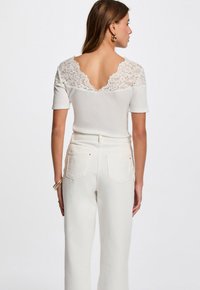 Cremefarvet top med korte ærmer og blomsterdækkende lace bagdetalje, med v-udskæring; sammen med højtaljede hvide jeans.