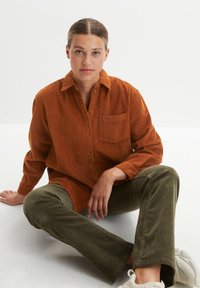 Chemise boutonnée en velours côtelé couleur rouille avec une poche poitrine ; associée à un pantalon en velours côtelé vert olive et des baskets blanches.