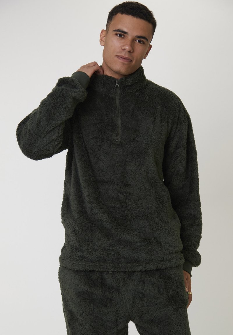 Ung man klädd i en mörkgrön, fluffig zip-up-tröja och matchande byxor, stående mot en enkel ljus bakgrund med ett avslappnat uttryck.