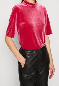 Top en velours à manches courtes de couleur rose vif avec un col montant. Porté avec un pantalon en cuir noir, affichant une texture lisse et un design ajusté.