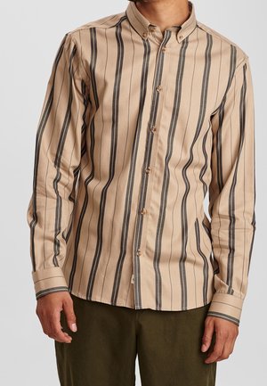 Mężczyzna w beżowej koszuli na guziki z pionowymi czarnymi paskami i oliwkowozielonych spodniach, stojący na tle jednolitego jasnego tła.