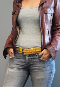 Braune Lederjacke, graues Tanktop, gelber Gürtel und ausgewaschene graue Jeans. Hände in den Taschen; einfaches, lässiges Outfit.