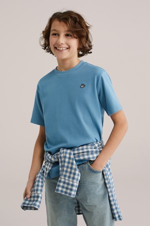 Smaidīgs bērns ar cirtainiem matiem, tērpies zilā T-kreklā un gaišās džinsās, ar zilu un baltu rūtainu kreklu sasietu apkārt viduklim.