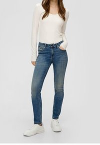 Light beige long-sleeve shirt paired with fitted blue denim jeans. Jeans feature a front pocket and slight fading. White sneakers complete the outfit.