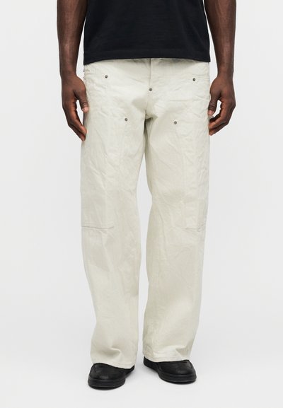 Pantalones cargo de color beige claro con un ajuste relajado, que cuentan con múltiples bolsillos, textura sutil y remaches metálicos. Combinados con zapatos negros.