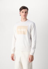 Weißer Pullover aus weichem Stoff, mit einem großen beigen rechteckigen Logo, das das Wort "HUGO" in Weiß zeigt. Gerippte Bündchen und Saum.