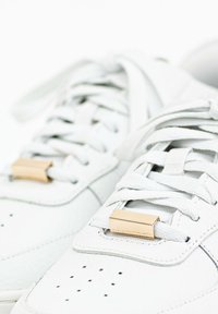 White leather sneakers featuring flat laces secured with gold-tone accents. Textured surface with perforations and stitching details.