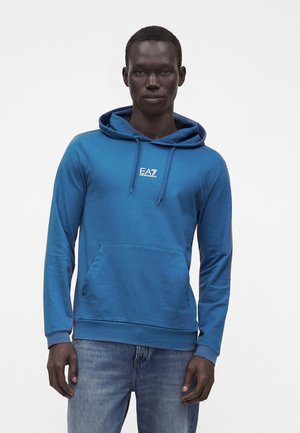 Άνδρας που φοράει μπλε φούτερ EA7 Emporio Armani με μπροστινό τσέπη και κορδόνια, συνδυασμένο με μπλε τζιν, στέκεται μπροστά σε λευκό φόντο.