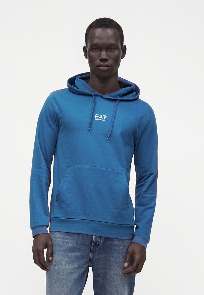 Άνδρας που φοράει μπλε φούτερ EA7 Emporio Armani με μπροστινό τσέπη και κορδόνια, συνδυασμένο με μπλε τζιν, στέκεται μπροστά σε λευκό φόντο.