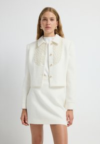 Chaqueta corta de bouclé blanca con cuello adornado con perlas, con un cierre de botones en el frente y hombros estructurados, combinada con una falda a juego.