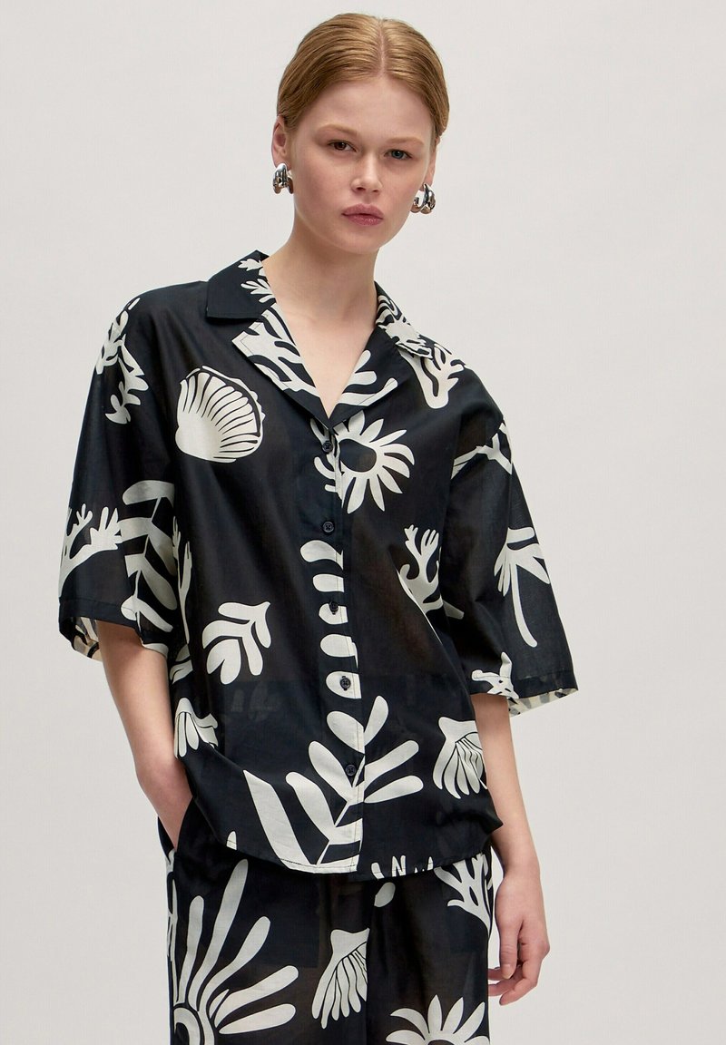 Schwarzes Kurzarmhemd mit einem weißen Blumen- und Muschelmuster, Knopfverschluss und Kragen. Dazu passende Hose.