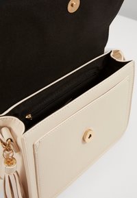 Béžová kabelka z umělé kůže se zlatým zapínáním na patent, černou textilní podšívkou a malou vnitřní kapsou na zip. Součástí je ozdobný střapec.