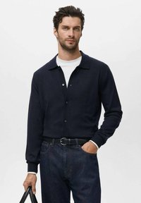 Cárdigan azul marino oscuro con cuello tipo solapa, cierre de botones en la parte delantera y puños acanalados. Combinado con una camisa blanca y jeans azul oscuro.