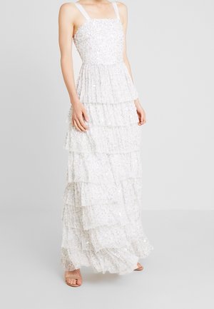 Frau in einem ärmellosen weißen Stufenkleid mit Paillettenverzierungen und quadratischem Ausschnitt, die vor einem schlichten hellen Hintergrund steht.