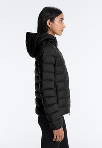 Fekete kapucnis steppelt dzsekit viselő nő profilból, egyszínű világos háttér előtt áll.