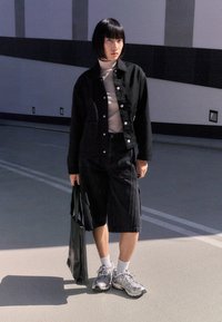 Schwarze Jeansjacke mit Knopfverschluss, kombiniert mit einem schwarzen Rollkragenpullover und knielangen Shorts. Eine schwarze Tragetasche und graue Sneaker runden den Look ab.