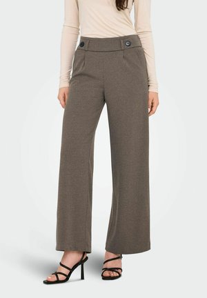 Vrouw in hoge, bruine broek met brede pijpen en knoopdetails, beige langemouwen top en zwarte gespsandalen met hakken.