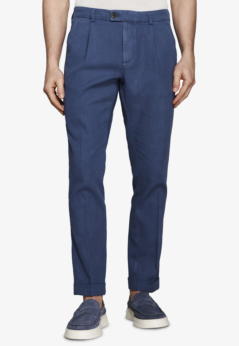 Hombre vistiendo pantalones azul ajustados con bajos doblados y zapatos deportivos azules a juego con suelas blancas.