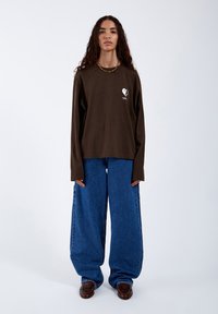Braunes Langarmshirt mit einem weißen Herz-Graphic, kombiniert mit weiten blauen Denim-Jeans. Weicher Stoff und lockere Passform.