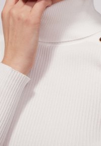 Prążkowany biały sweter z wysokim kołnierzem. Posiada gładką teksturę oraz pojedynczy złoty guzik w okolicy szyi.