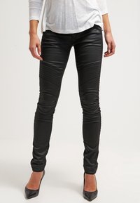 Pantalones ajustados de cuero sintético negro con detalles acanalados en los muslos y acabado elegante, combinados con zapatos de tacón alto negros.