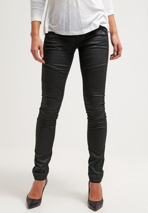 Pantalons skinny en faux cuir noir avec des détails nervurés sur les cuisses et une finition lisse, associés à des chaussures à talons hauts noires.
