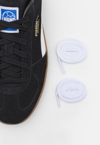 Schwarze Wildledersportschuhe mit weißen Akzenten und schwarzen Schnürsenkeln, versehen mit einer Gummisohle. Neben dem Schuh liegen zwei weiße Schnürsenkel.