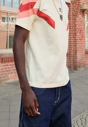 Mann trägt ein cremefarbenes Adidas-T-Shirt mit roten diagonalen Streifen und Löwenemblem, kombiniert mit dunkelblauen Jeans, steht auf einem Bürgersteig in der Stadt.