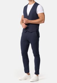 Gilet bleu marine avec un col net et deux boutons, associé à un pantalon sur mesure et des baskets blanches ; texture de tissu lisse, design minimaliste.