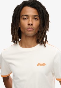 Cremefarvet bomulds T-shirt med orange detaljer på ærmerne og broderet "Superdry" logo på brystet; rund hals design.