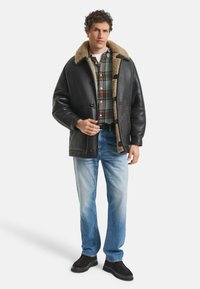 Zwarte leren jas met een beige schapenvachtkraag, grote knopen en een geruit overhemd daaronder, gecombineerd met lichtblauwe jeans en donkere schoenen.