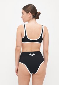 Juodas bikini komplektas, kuriame yra aukštai iškeltas dugnas ir trumpas viršus, abu papuošti baltais apvadais ir deimanto logotipu nugaros pusėje. Sklandžios tekstūros.