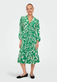 Abito floreale verde con maniche lunghe, scollatura a V e design a gonna a strati. Indossato con ballerine nere con dettagli in perle.