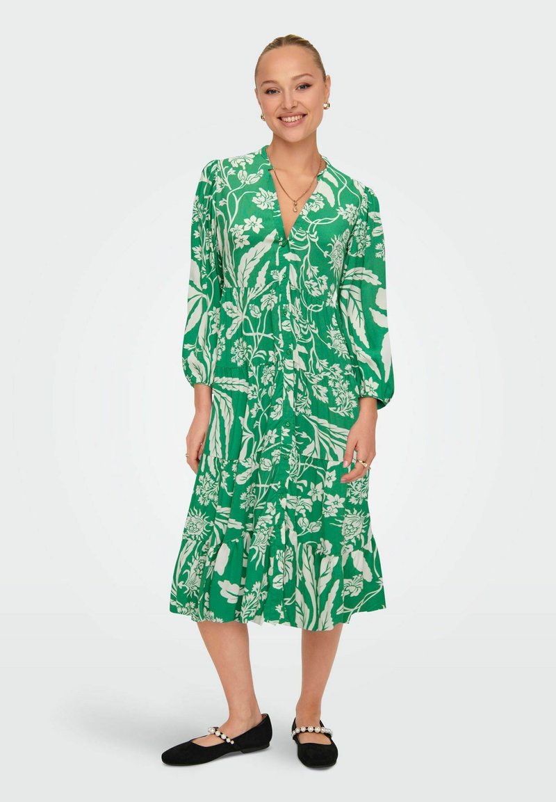 Abito floreale verde con maniche lunghe, scollatura a V e design a gonna a strati. Indossato con ballerine nere con dettagli in perle.