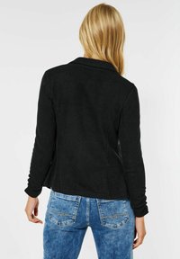 Musta sovitettu takki, jossa on kaulus, pitkät hihat ja teksturoitu kangas, yhdistettynä sinisiin farkkuihin, joissa on ommeltuja koristeita takataskuissa.