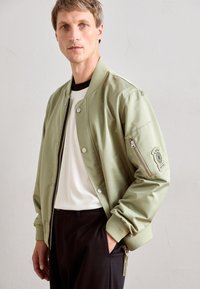 Man wearing light green bomber jacket over white ribbed shirt and black pants, standing against plain light wall background.