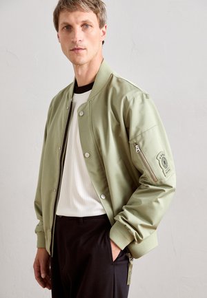 Homme portant une veste bomber verte claire par-dessus une chemise blanche à côtes et un pantalon noir, se tenant devant un fond mural clair et uni.