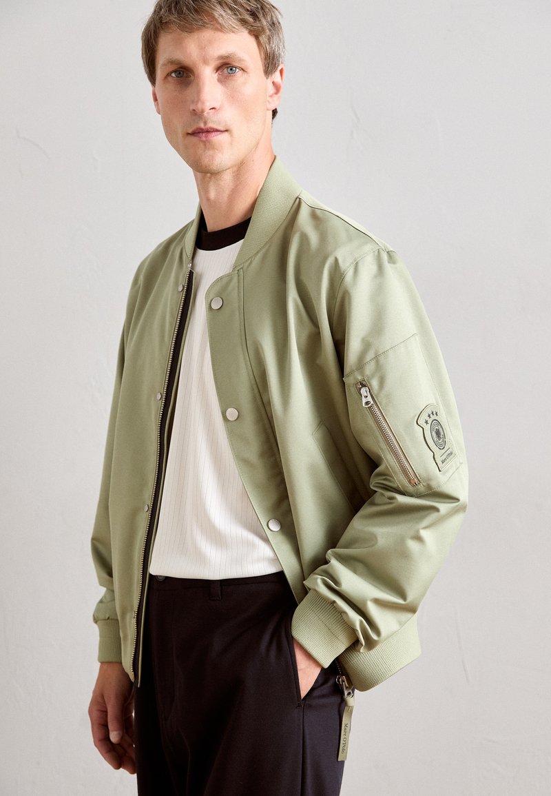 Man wearing light green bomber jacket over white ribbed shirt and black pants, standing against plain light wall background.