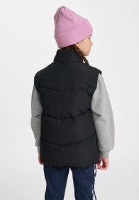 Schwarze Steppweste mit hohem Kragen und gestepptem Muster, getragen über einem grauen Sweatshirt, kombiniert mit marineblauen Jogginghosen und einer pinken Mütze.
